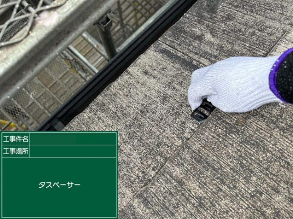 スレート屋根の端部にタスペーサーを挿入している作業の様子。足場が見える位置から撮影
